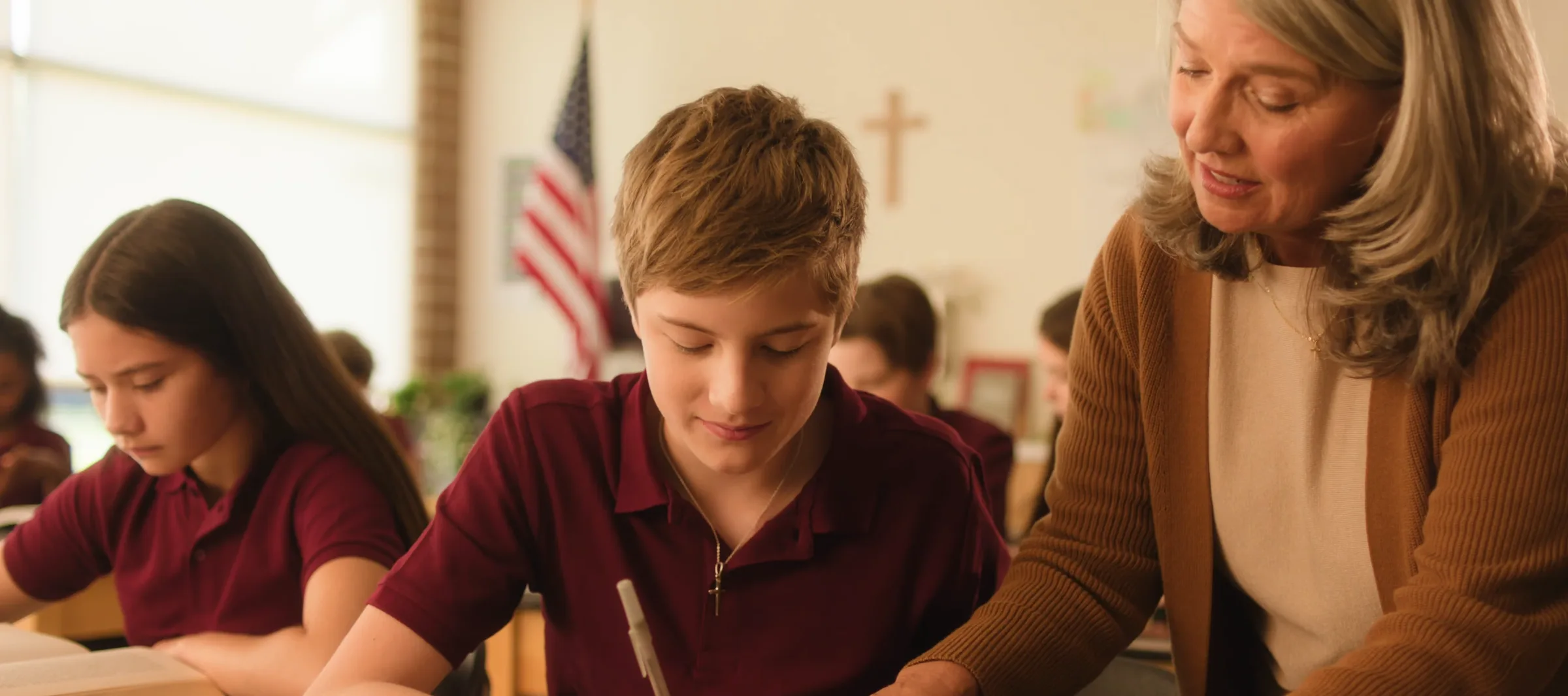 A boy in a maroon school shirt writing at his desk while his teacher leans in to help